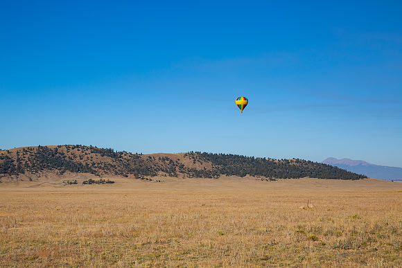 15.5 MB, 5172 x 3448: A rainbow colored hot air ballon floats above a valley and mountains.