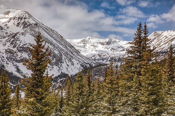 27.4 MB, 6717 x 4478: Snow covered mountain peaks and green pine trees under partly cloudy skies