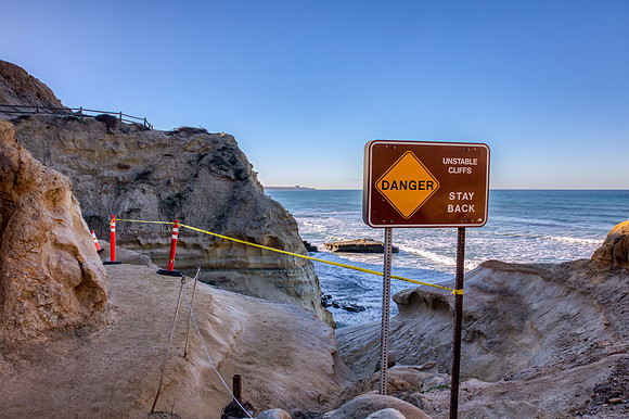 13.6 MB, 6300 x 4193: Danger sign, yellow tape, and orange cones warns hikers of unstable cliffs on a trail down to the Pacific ocean.