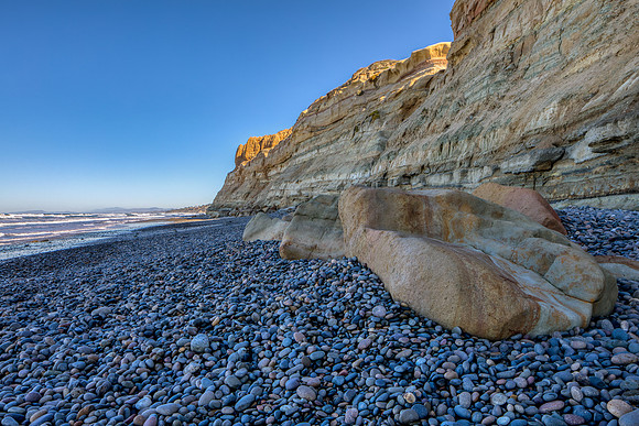 24.0 MB, 6720 x 4480: Beach covered in brown, blue and tan smooth rocks as sunlight illuminates the brown cliff tops.
